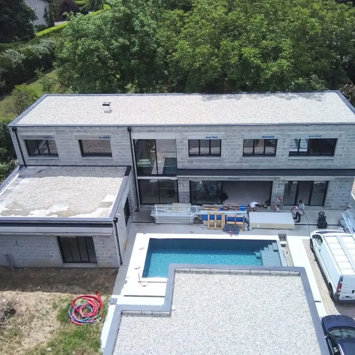 Maison achevée avec toiture-terrasse et grandes baies, sa piscine et ses plages, entourée d'arbres.
